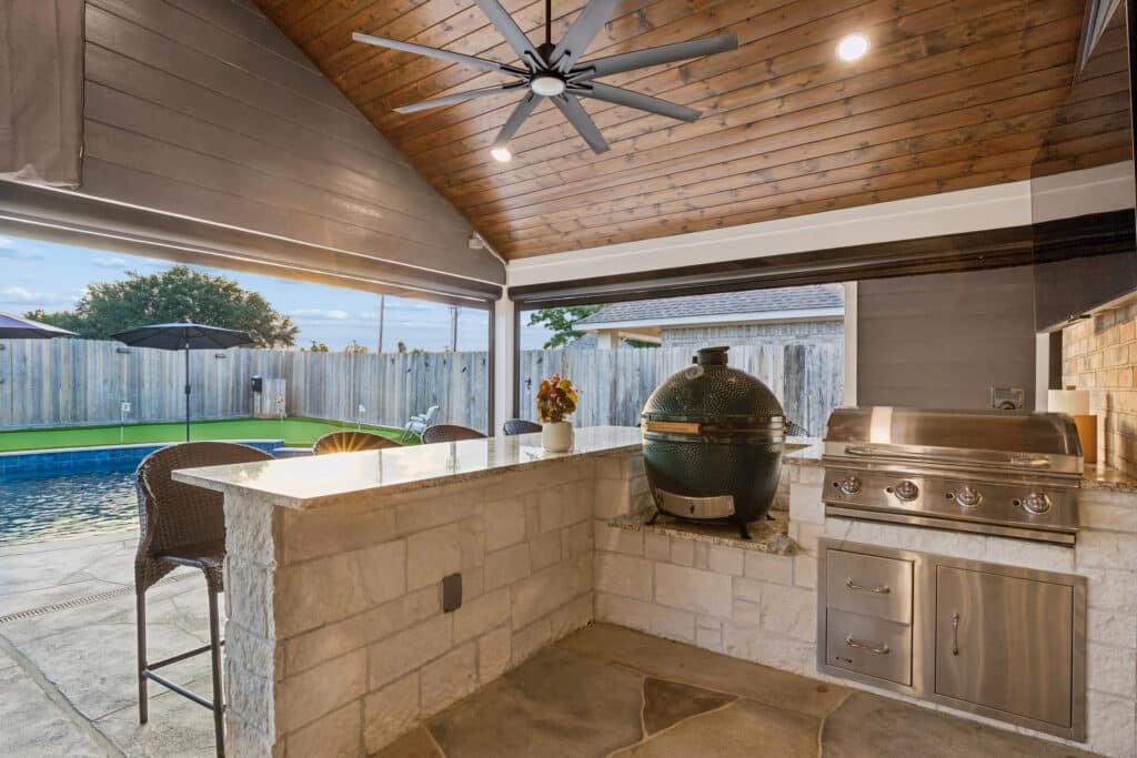 Outdoor kitchen under a wooden pergola with a stone bar, a green ceramic smoker, and a stainless grill by a backyard pool.