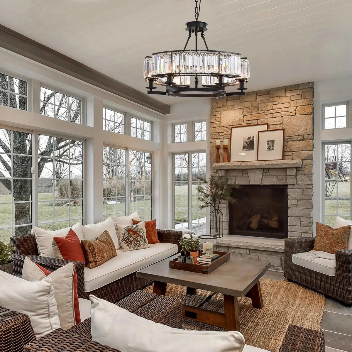Bright sunlit living room with a stone fireplace, wicker sofas and chairs, white cushions, and orange accent pillows around a rectangular coffee table in the center.